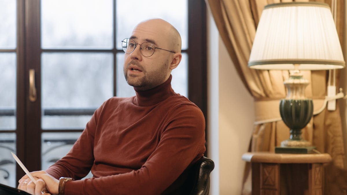 Bald man in glasses speaking while seated indoors, with a warm lamp and curtains in the background.